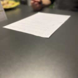 Eine Seite Papier liegt auf einem dunklen Tisch. Im hintergrund kann man angedeutet Menschen erkennen. Symbolbild.